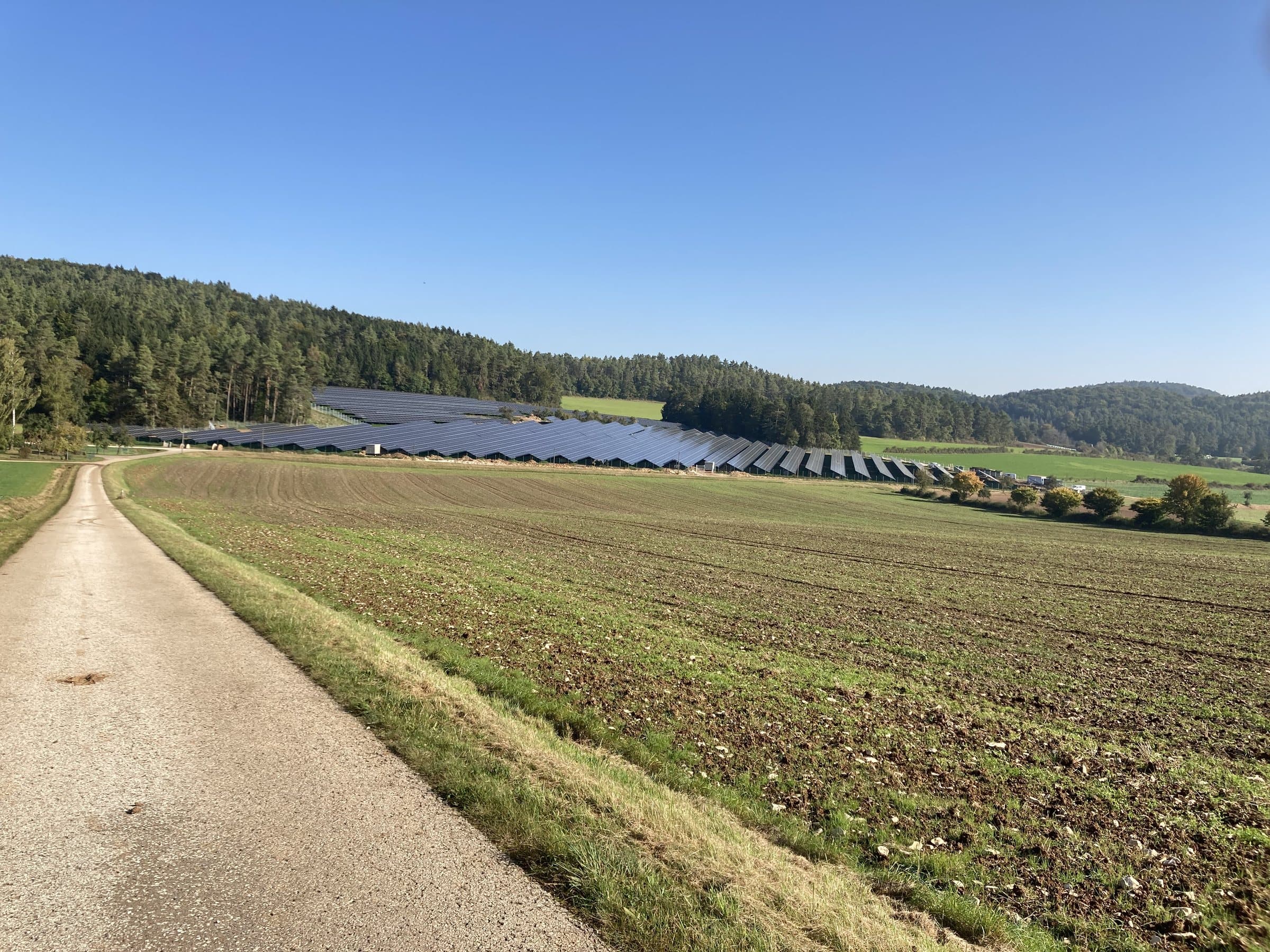 Blick über einen Feldweg auf die Freiflächenanlage PV Oberfeld am Waldrand in hügeliger Landschaft
