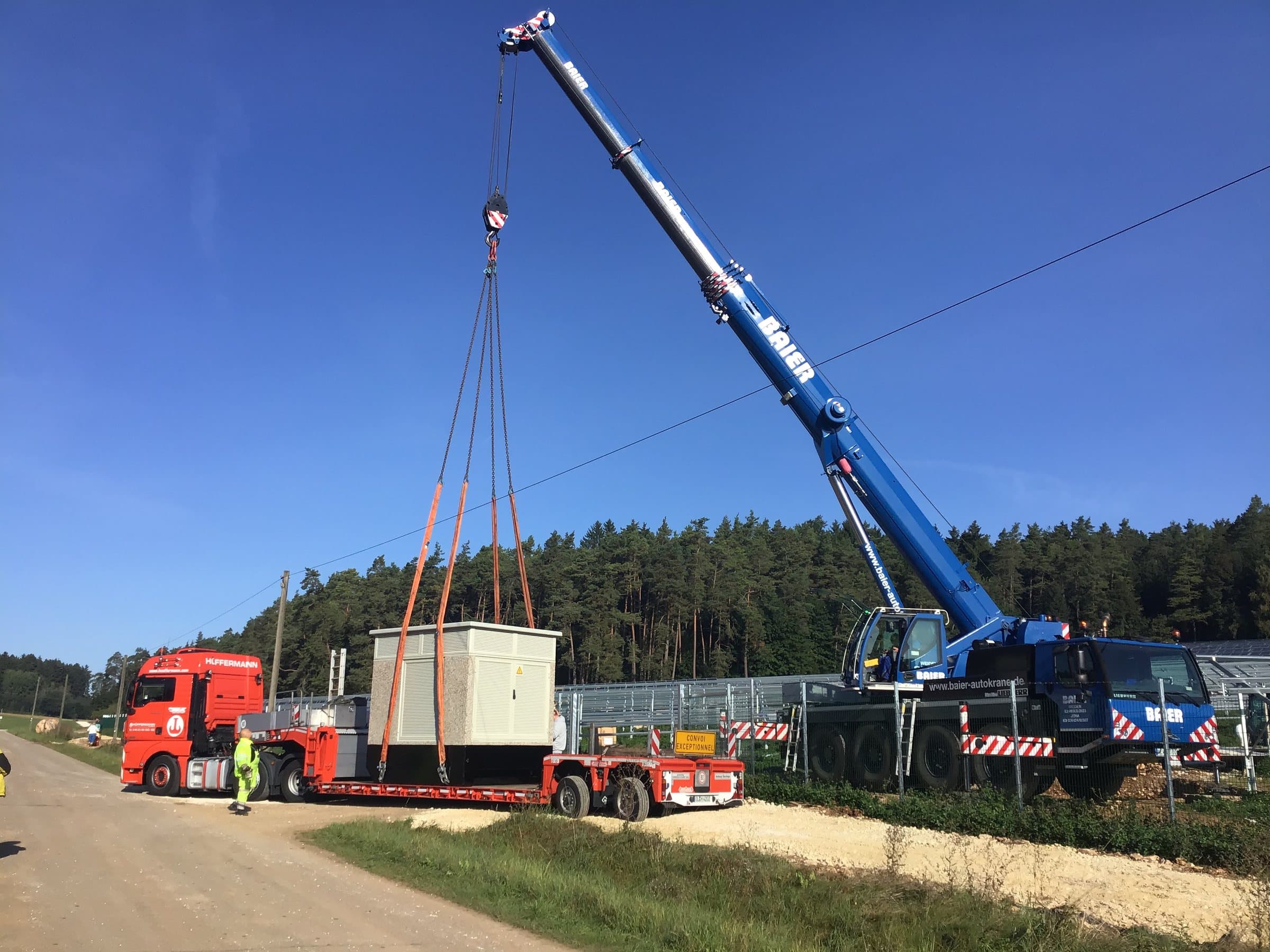 Ein Autokran hebt die Trafostation vom Tieflader auf ihren Platz am Zaun der PV-Anlage Oberfeld