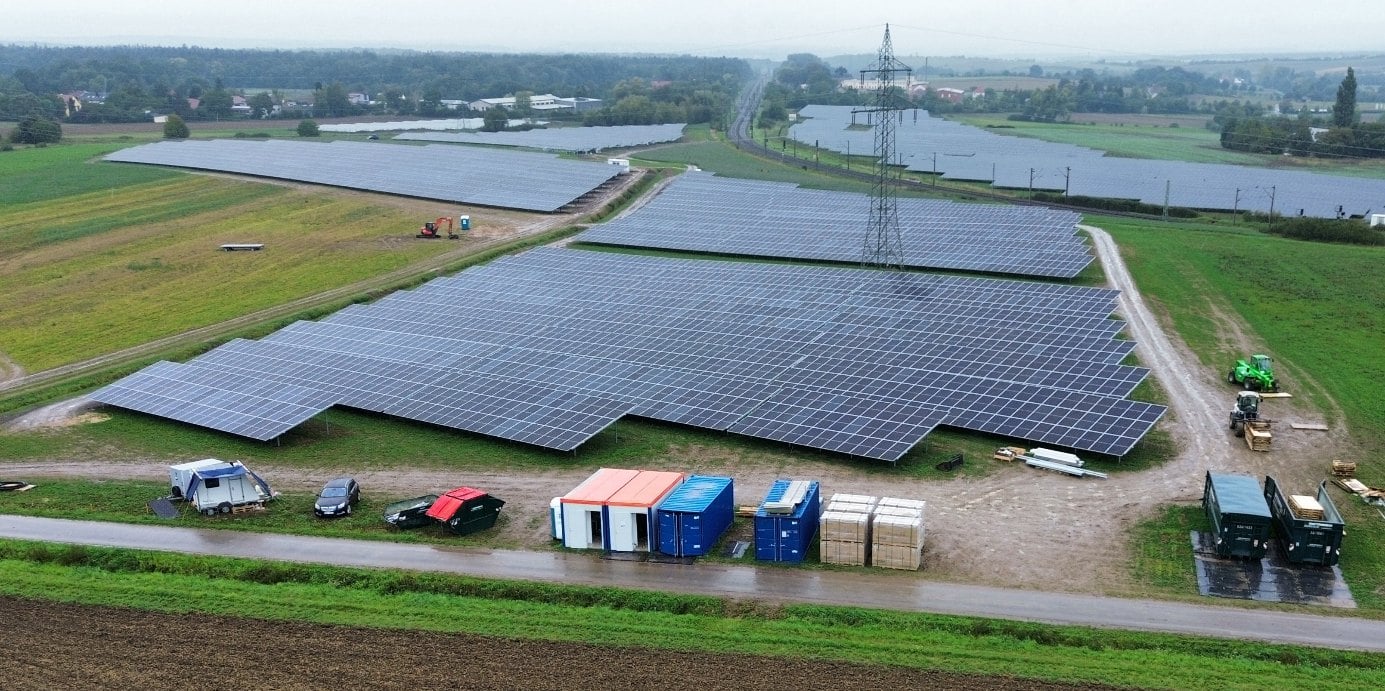 Luftaufnahme des neu errichteten Solarparks bei Kitzingen mit Baucontainern am Feldweg, darüber eine Hochspannungsleitung