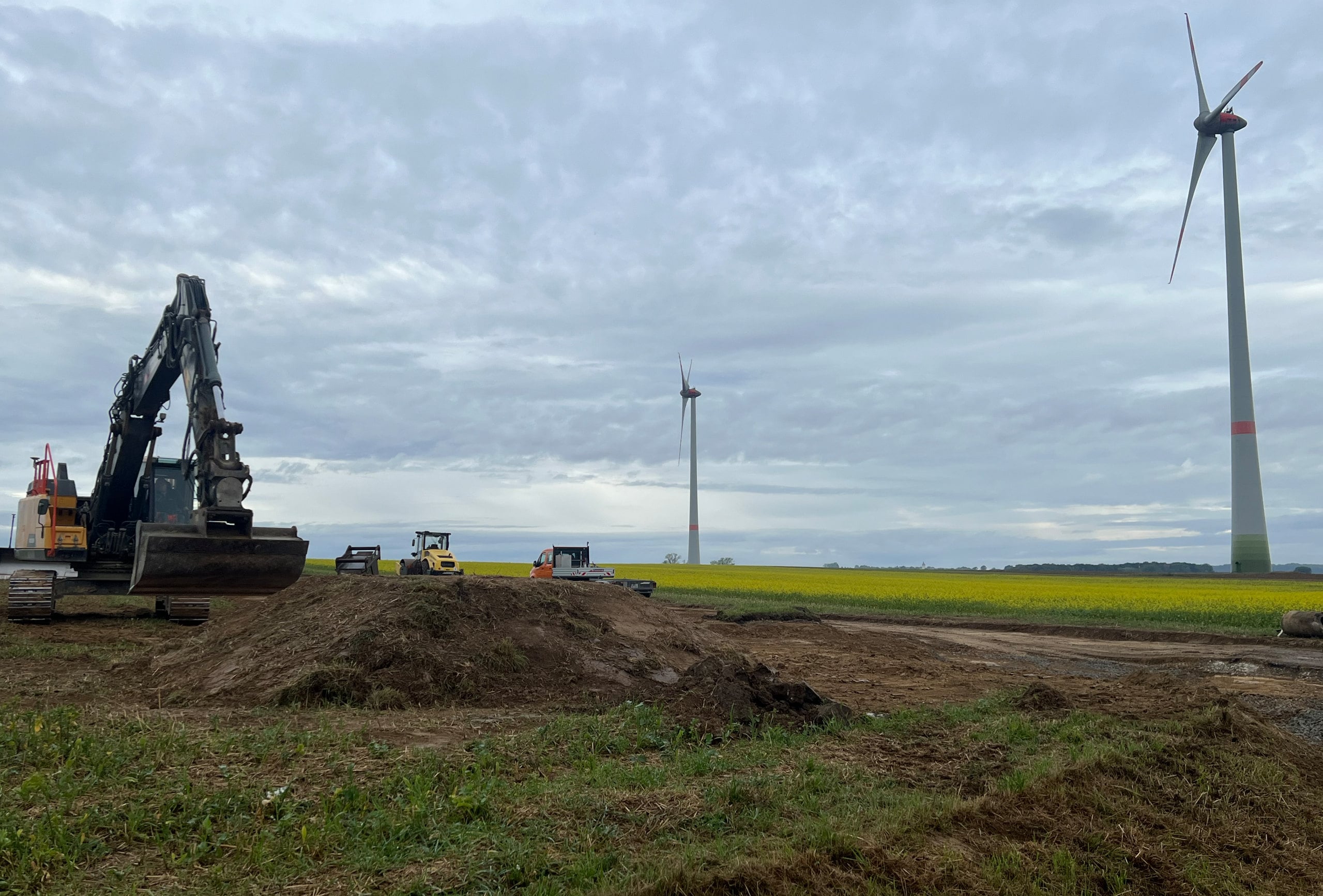 Bagger auf der Baustelle in Insingen, im Hintergrund drehen sich Windenergieanlagen über einem Rapsfeld